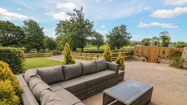 Un espace patio aménagé avec un canapé en osier et une table, entouré de verdure et délimité par une clôture en bois.