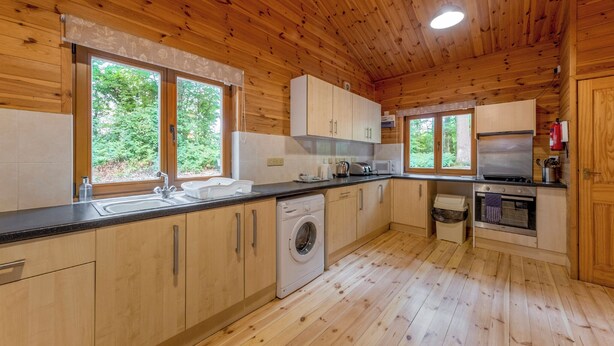 A wooden cabin kitchen with a washing machine, oven, and large windows. - Spacious and comfortable lodge with contemporary styling. (Ellesmere)