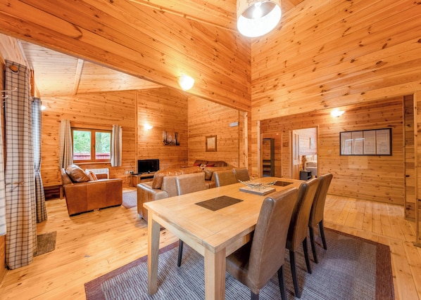 A wooden cabin interior with a dining table, chairs, a sofa, a TV, and a window. - Spacious and comfortable lodge with contemporary styling. (Ellesmere)