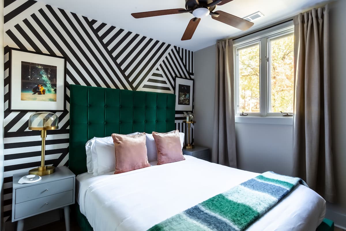Habitación de hotel con cabecera verde acolchada, un ventanal con cortinas, cama con ropa de cama blanca y una manta a rayas, mesita de noche con lámpara y pared con estampado geométrico.