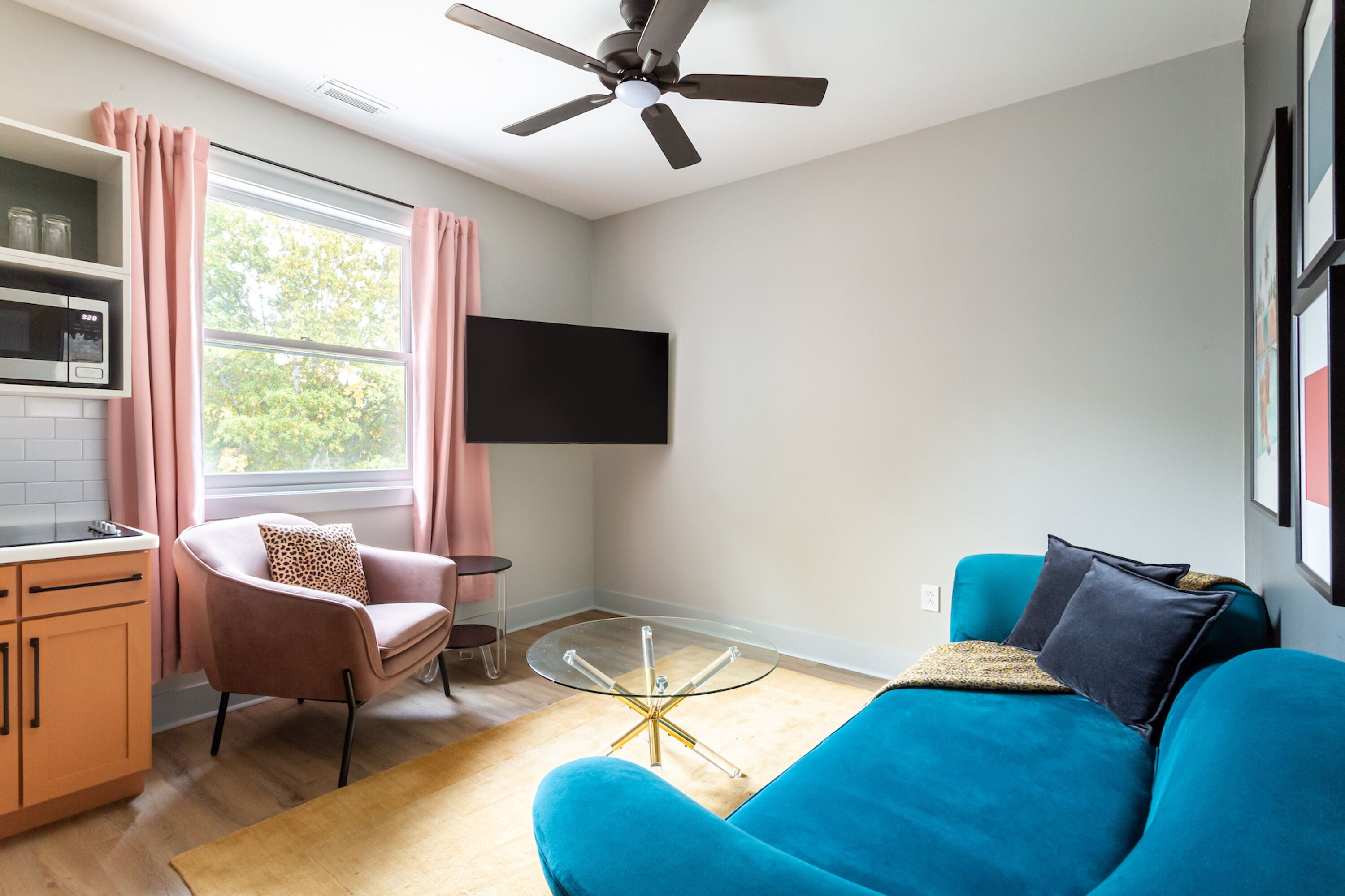 Una sala de estar moderna con un sofá color turquesa, un sillón rosa, una mesa de centro de vidrio, un ventilador de techo y un televisor de pantalla plana montado en la pared.