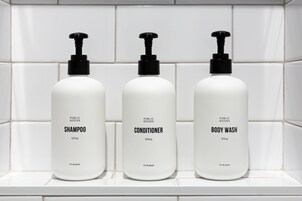 Three bottles of public goods shampoo, conditioner, and body wash on a white tiled shelf.