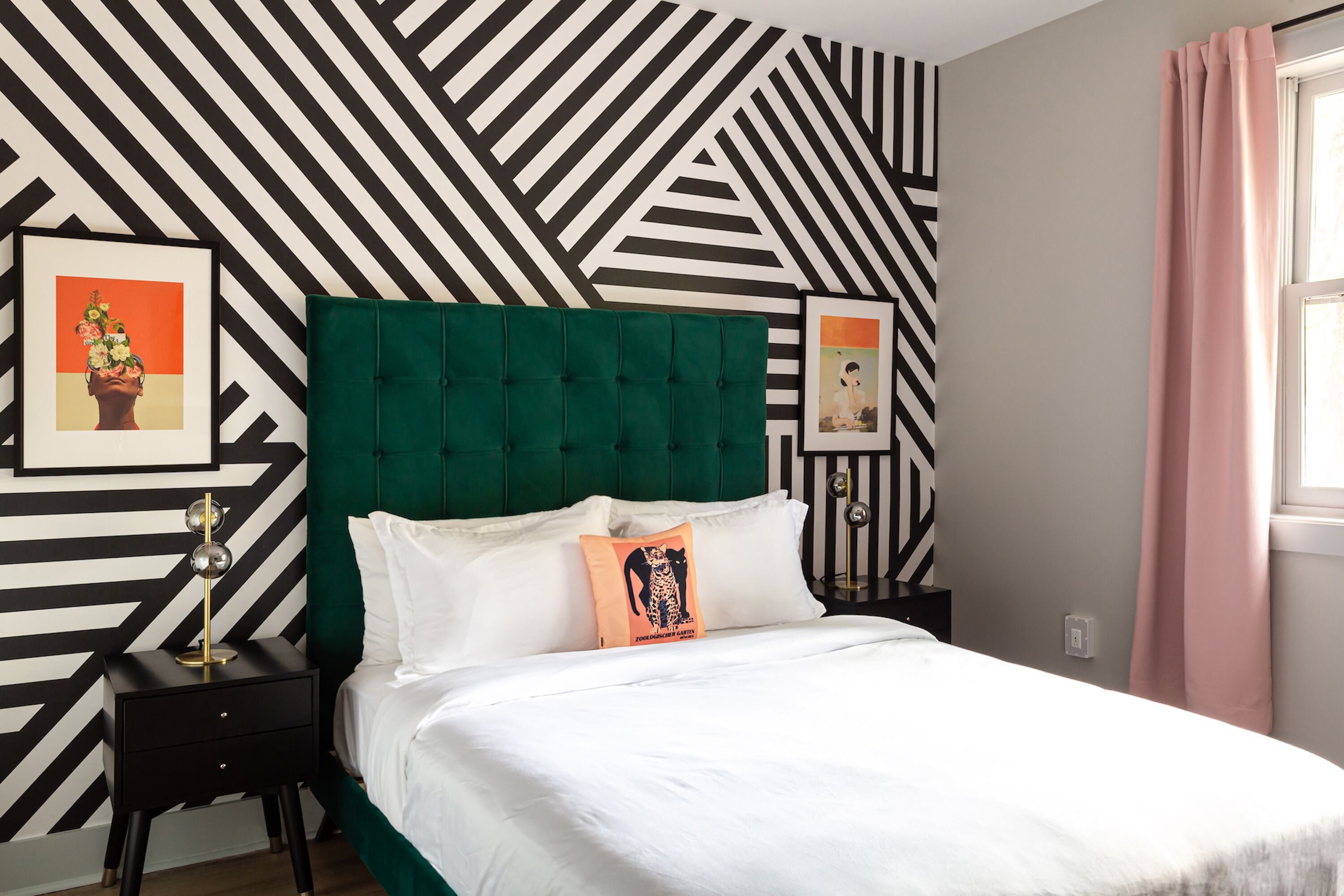 A bed with a green headboard, white bedding, and two pillows, one with a leopard print design. There are two bedside tables with lamps, and the wall features a black and white geometric pattern.