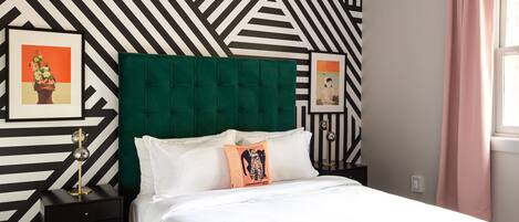 Una cama con cabecera verde, ropa de cama blanca y dos almohadas, una con estampado de leopardo. Hay dos mesitas de noche con lámparas, y la pared presenta un diseño geométrico en blanco y negro.