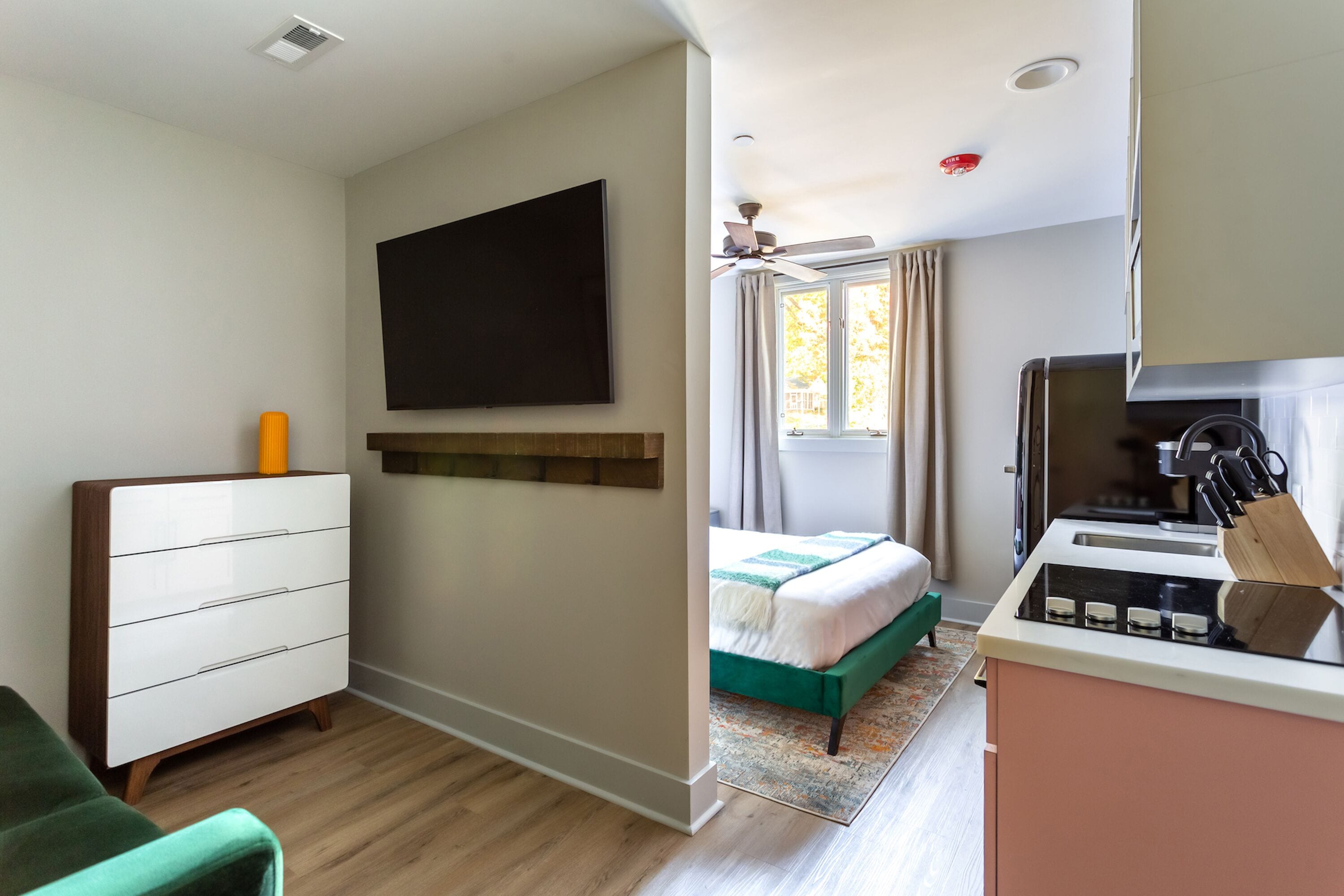 A modern hotel room with a flat-screen TV, a bed with a green accent, a kitchenette with a stove and knife block, and a ceiling fan.