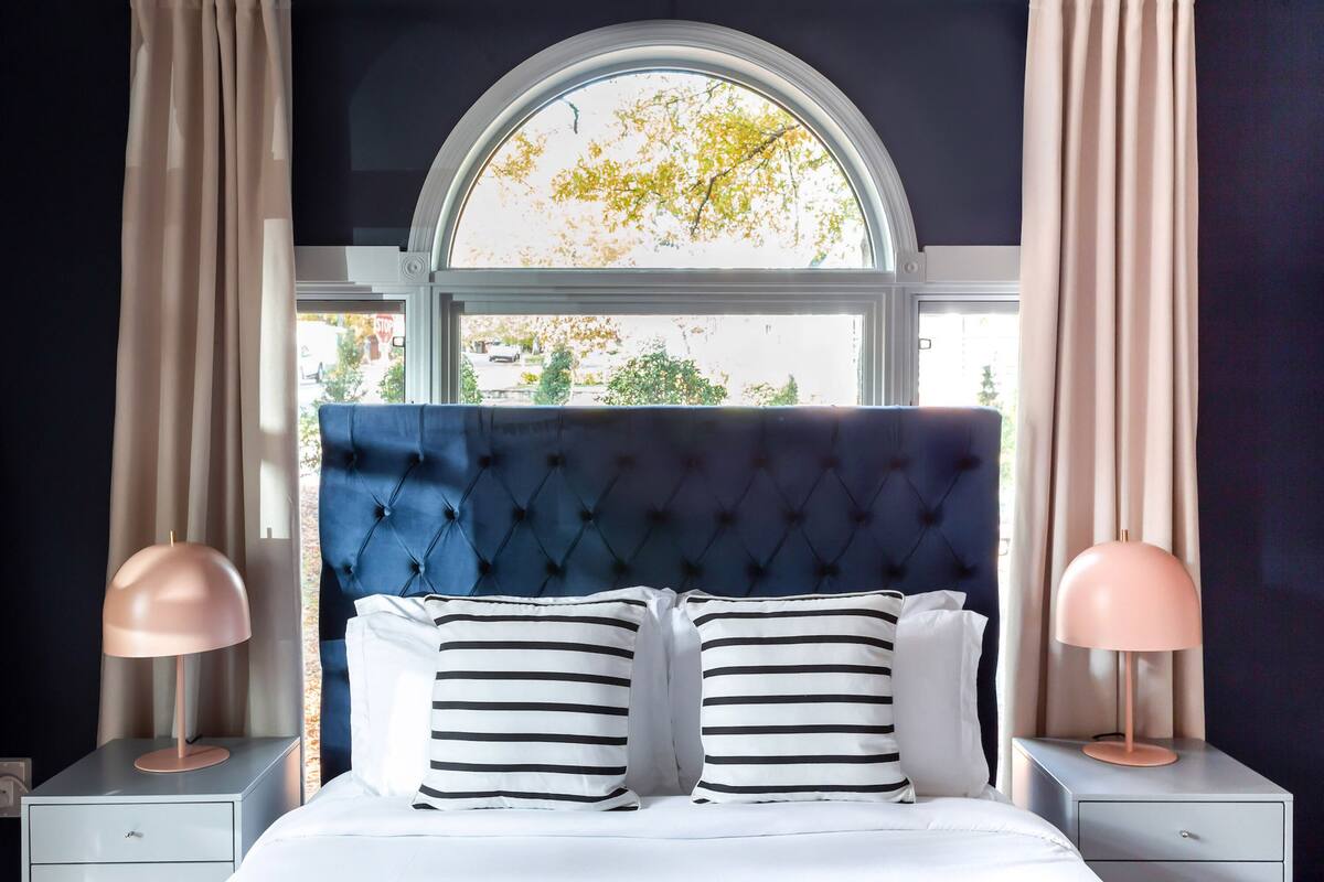Un dormitorio con cabecera capitonada, un ventanal con cortinas, dos mesitas de noche con lámparas y almohadas a rayas.