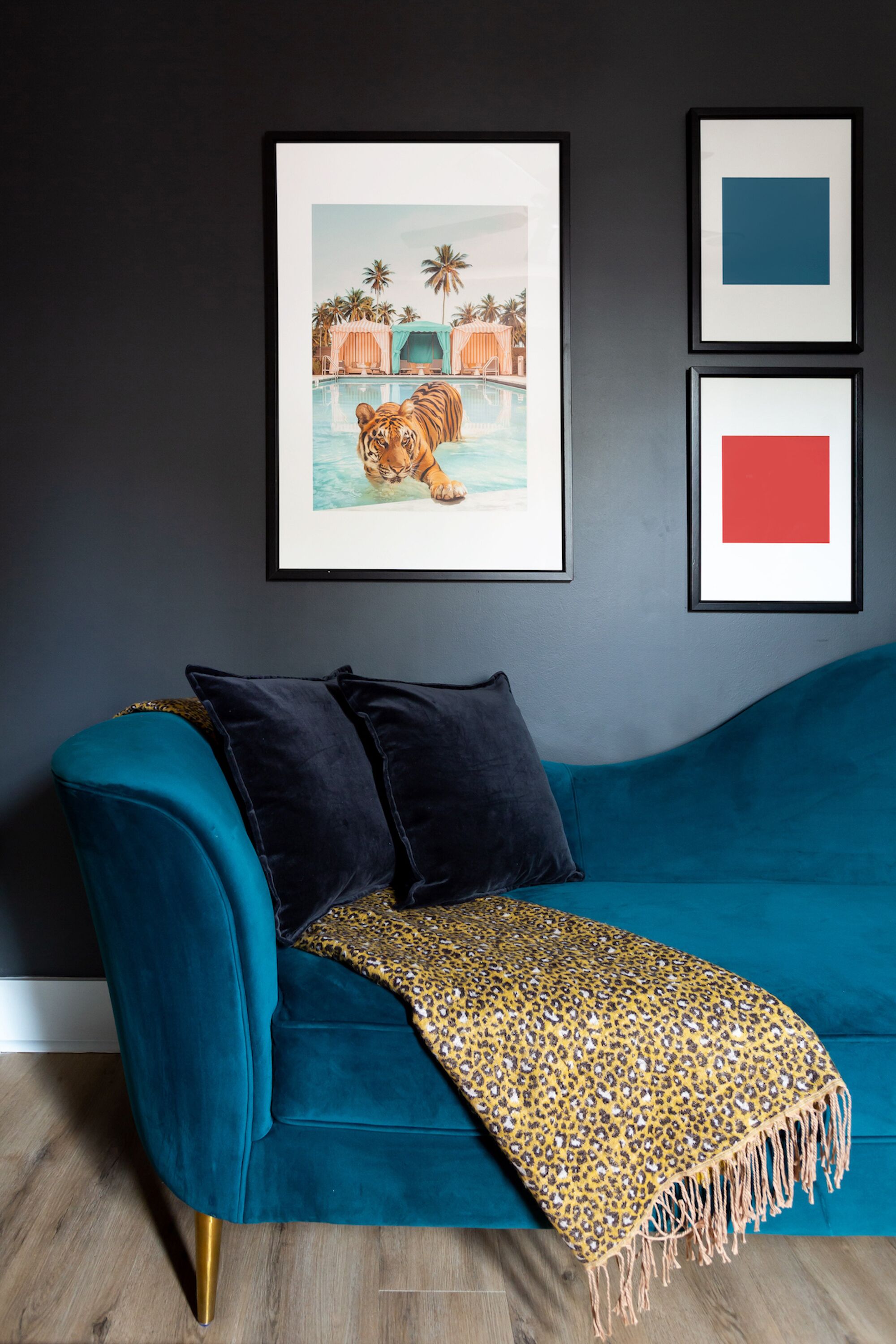 Un canapé de couleur turquoise agrémenté d’un plaid à motifs léopard, une illustration de tigre encadrée et deux formes géométriques encadrées sur un mur sombre.