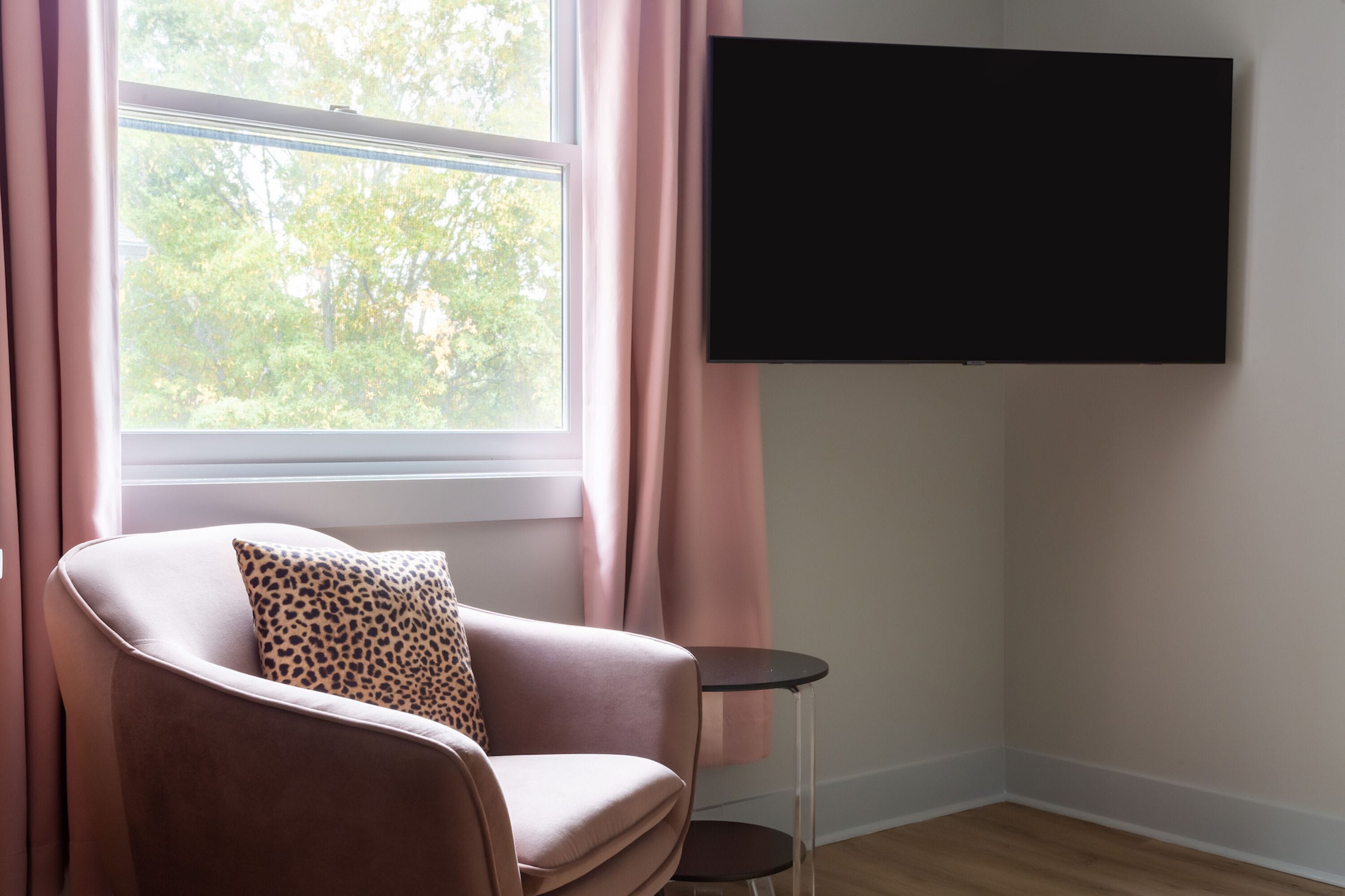A room with a pink armchair, a small round table, a TV mounted on the wall, and a window with curtains.