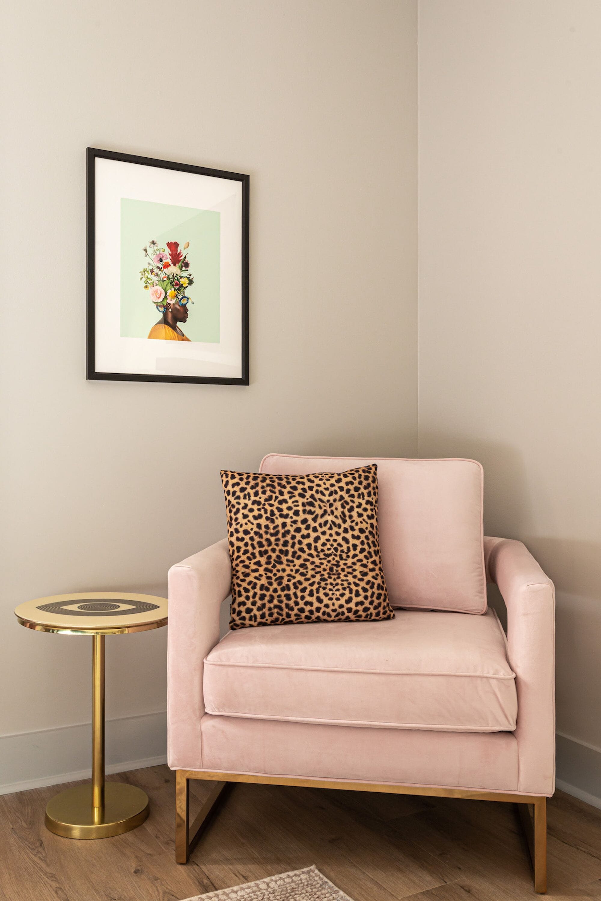 Un fauteuil rose agrémenté d’un coussin à motifs léopard, une petite table ronde avec un pied doré, et un tableau encadré représentant une composition florale accroché au mur.