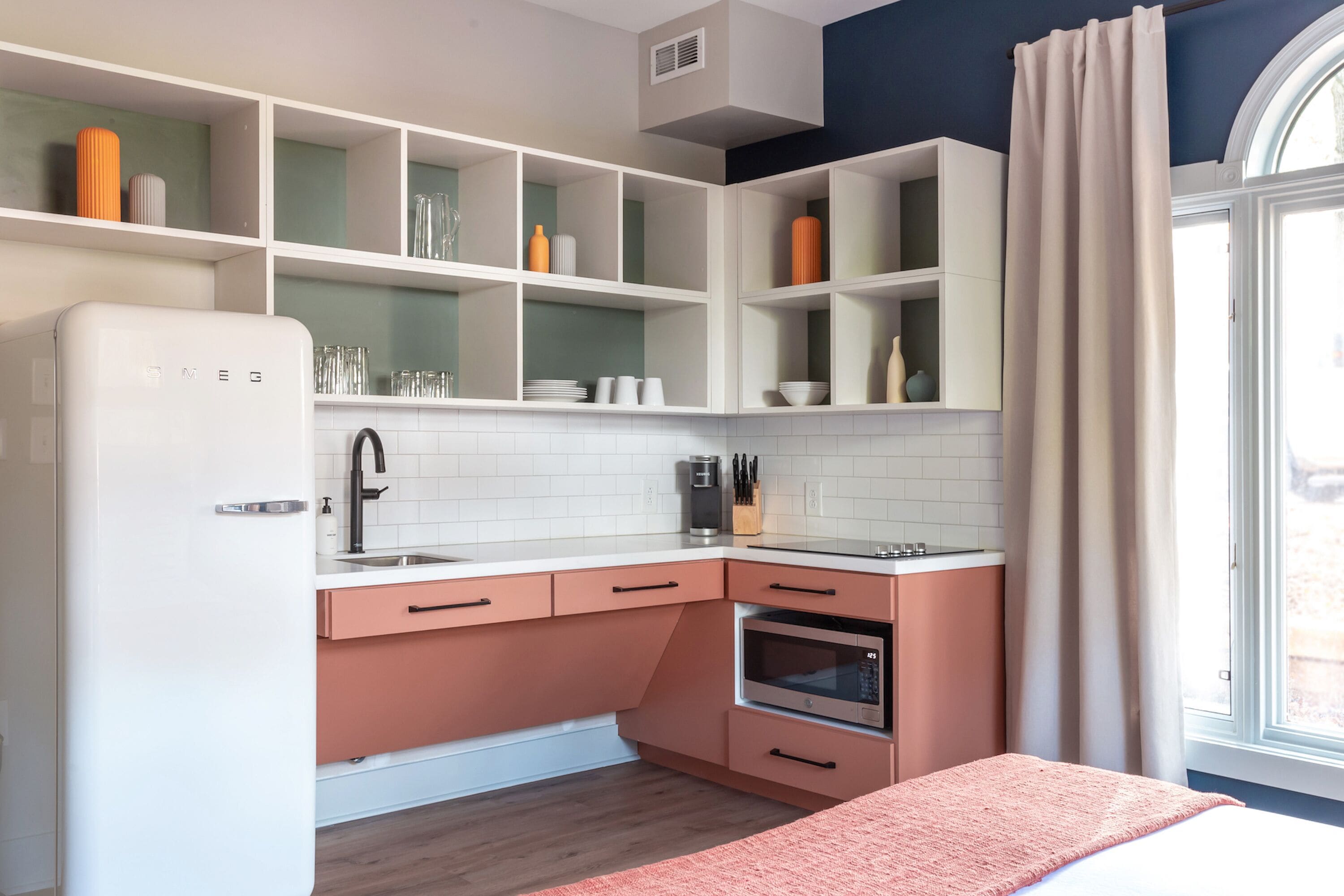 A compact kitchen with white and peach cabinetry, a built-in oven, and a window with curtains.