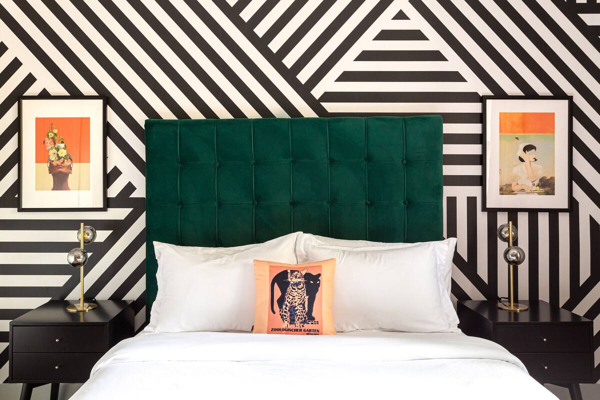 Una cama con cabecera tapizada en verde, ropa de cama blanca y un cojín con una ilustración de un gato. Hay dos mesitas de noche con lámparas y cuadros en la pared.