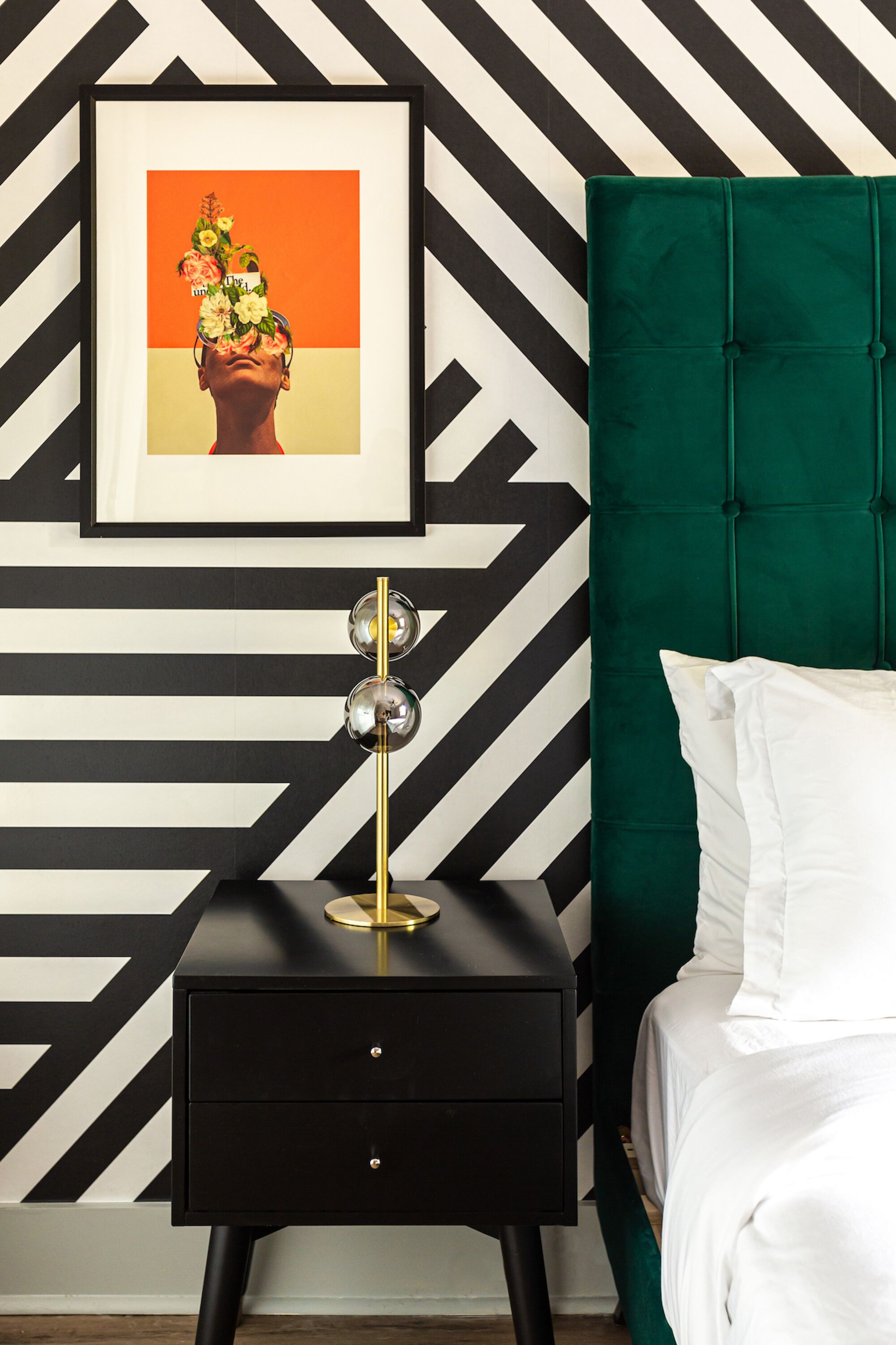 A modern bedroom with a black and white chevron-patterned wall, a green upholstered headboard, a bedside table with a gold lamp, and a framed picture above the bed.