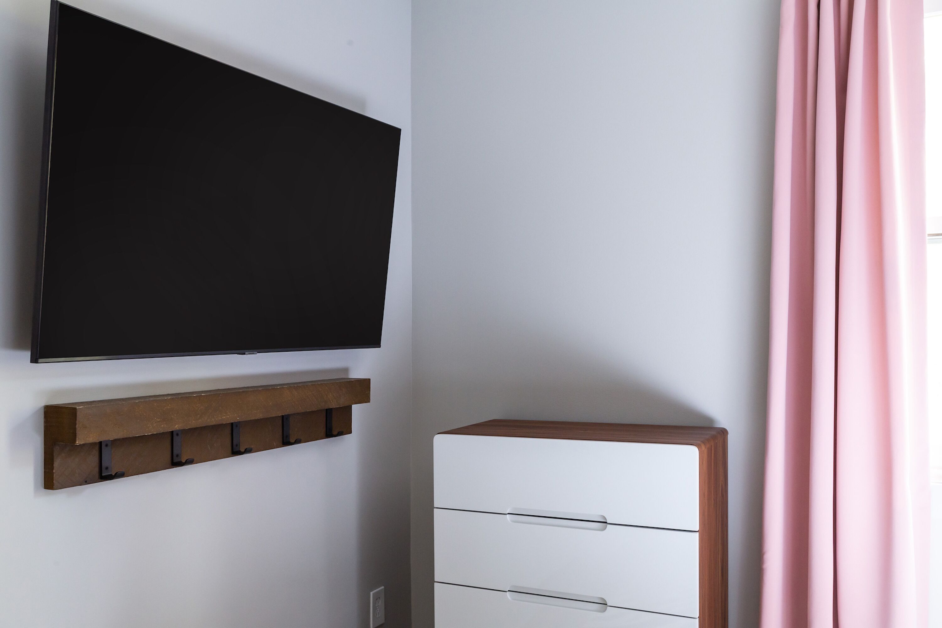 A room with a mounted flat-screen TV, a wooden shelf, a white dresser with drawers, and a pink curtain.