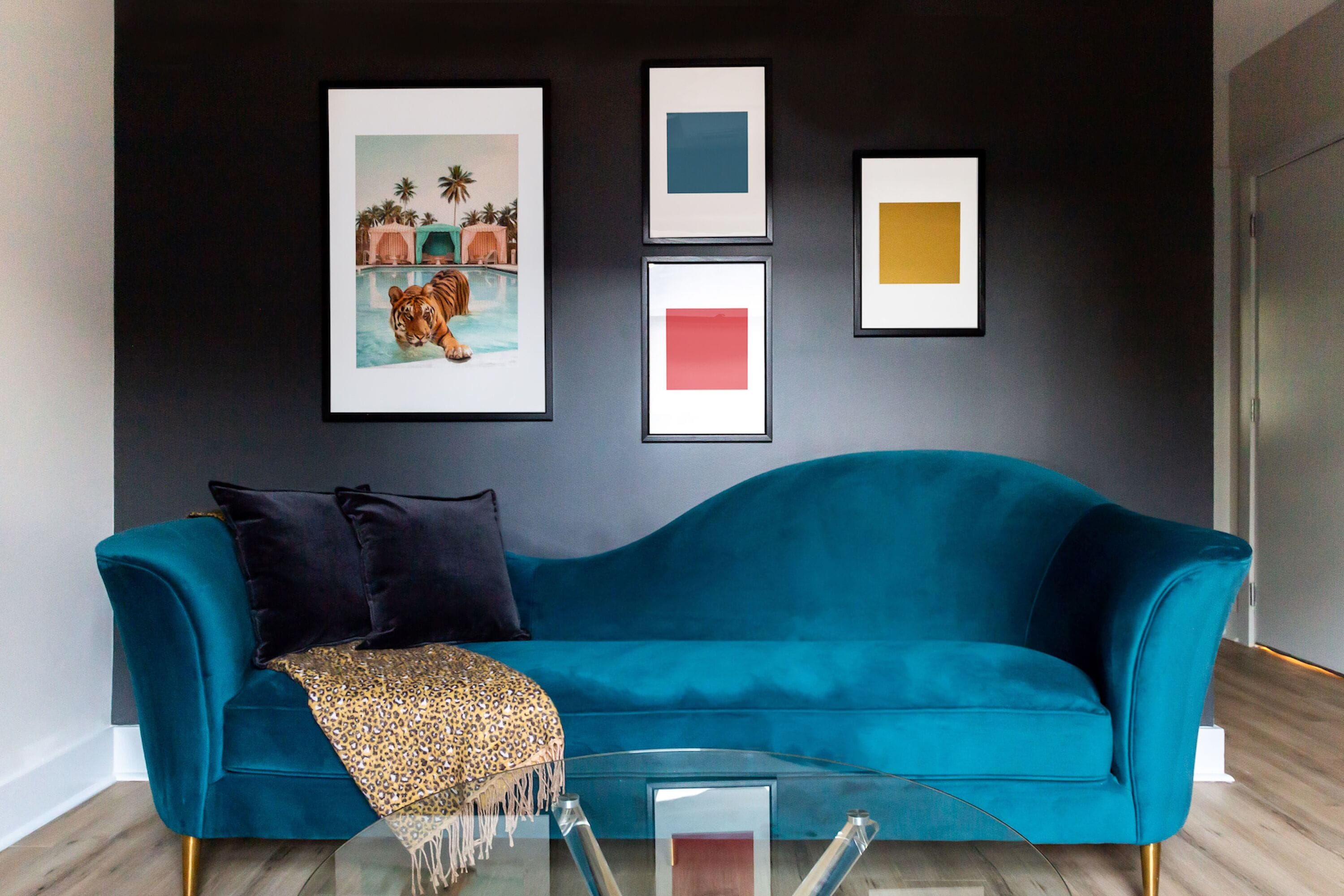 A teal sofa with a glass coffee table in a modern living room with a dark wall and framed artwork.