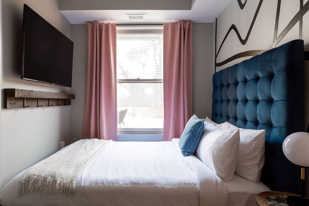 Un dormitorio ordenado con una cama grande, un televisor de pantalla plana montado en la pared y una ventana con cortinas rosadas.