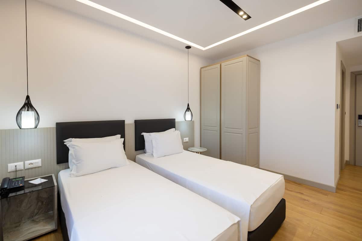 A modern hotel room with two single beds, a bedside table with a phone, a wardrobe, and a hanging light fixture.