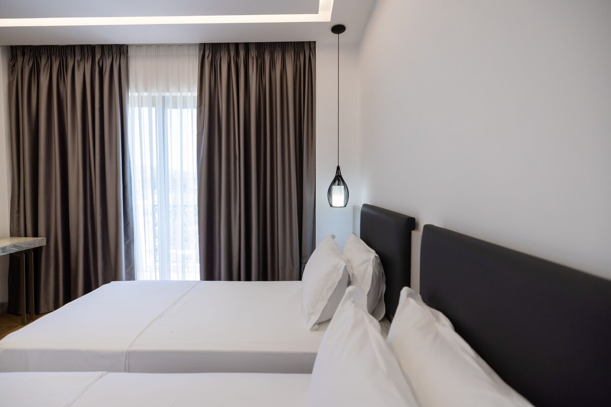 A hotel room with a bed, a bedside table, a hanging light fixture, and a window with curtains.