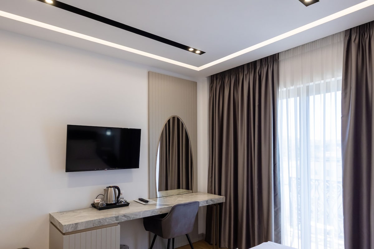 A hotel room with a bed, a desk with a flat-screen TV, a chair, and a window with curtains.
