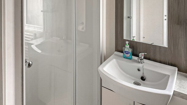 Ein Badezimmer mit Glasduschtür, einem weißen Waschbecken mit Spiegel und einer Flasche Carex Handseife.
