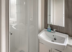 Un baño con mampara de vidrio, un lavamanos blanco con espejo y una botella de jabón de manos Carex.