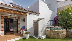 A modern outdoor area with a stone jacuzzi, a glass-enclosed shower, and a small seating area with a bench and potted plants.