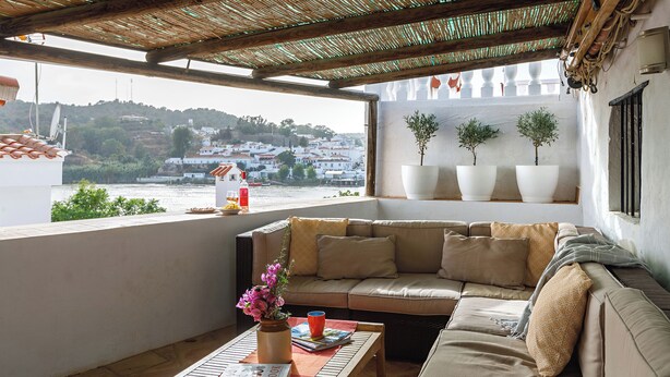 A balcony with a cushioned seating area, a table with a book and a vase, and a view of a town across a body of water.