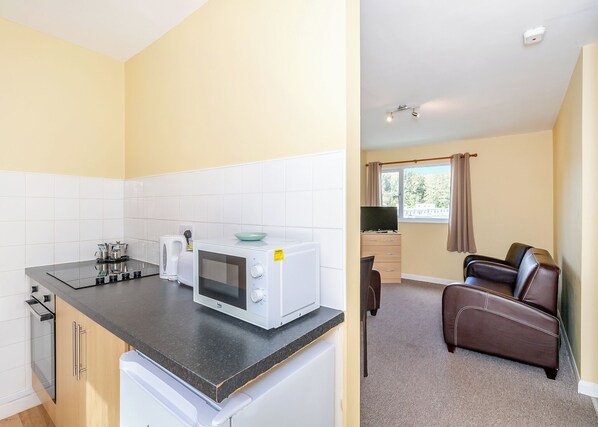 A compact kitchen with a microwave, kettle, and toaster, a dining area with a leather chair, and a window with curtains.