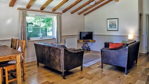 A cozy living room with a wooden ceiling, a television, two sofas, a dining table with chairs, and a window with curtains.