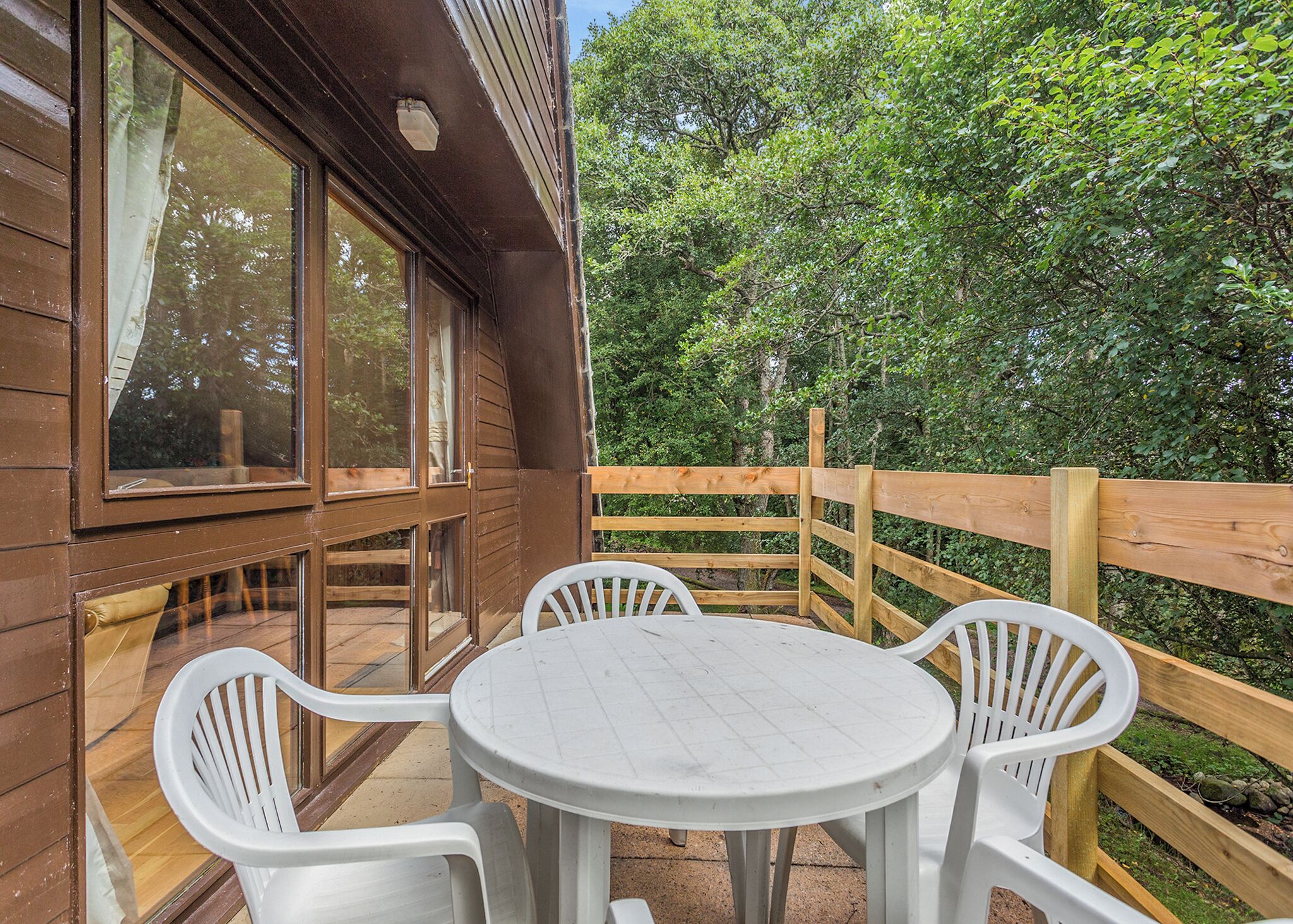 Een houten terras met een ronde tafel en vier stoelen, omgeven door een houten hek en bomen.