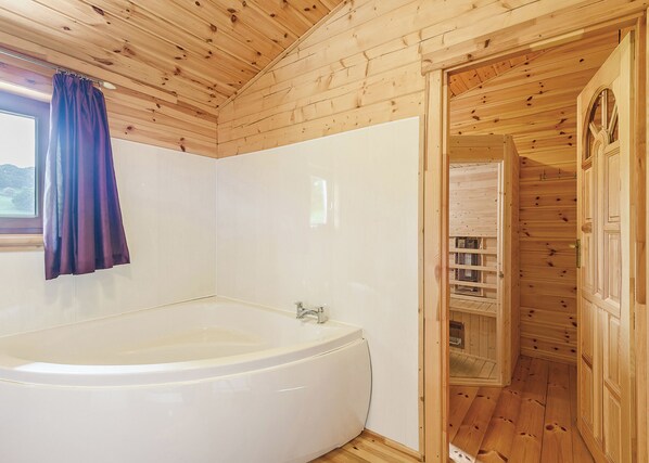 A wooden cabin interior with a large bathtub, a window with a curtain, and a wooden door leading to a closet.