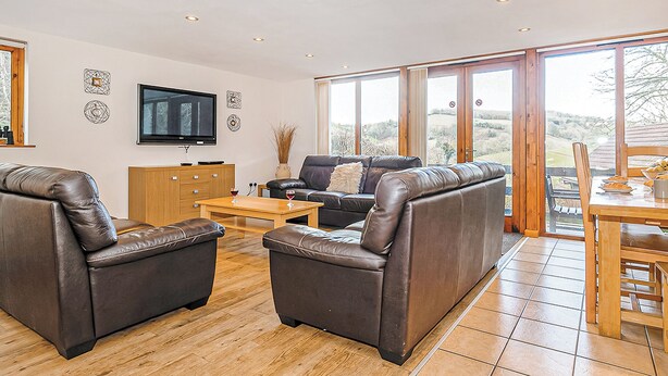 A spacious living room with leather sofas, a wooden coffee table, and a flat-screen TV mounted on the wall. - Luxury lodge residing at the top of the wooded valley. (Waterrow, Wiveliscombe)