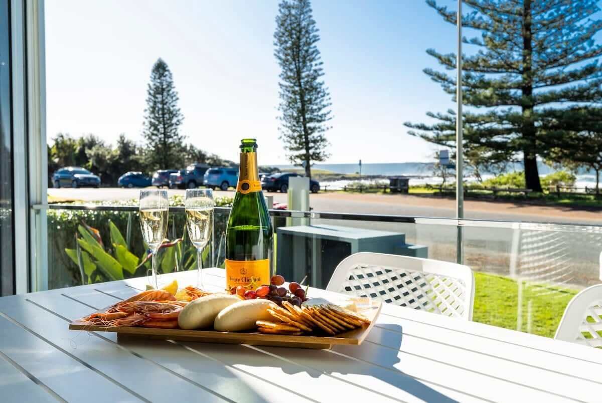A table with a bottle of champagne, glasses, and a platter of food including shrimp, bread, and crackers, set on a balcony with a view of the sea and parked cars.