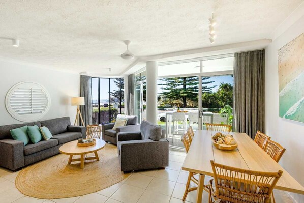 A spacious living room with a sofa, coffee table, and dining table set. - La Ronde One - Oceanstays - Unit 1 (Yamba)