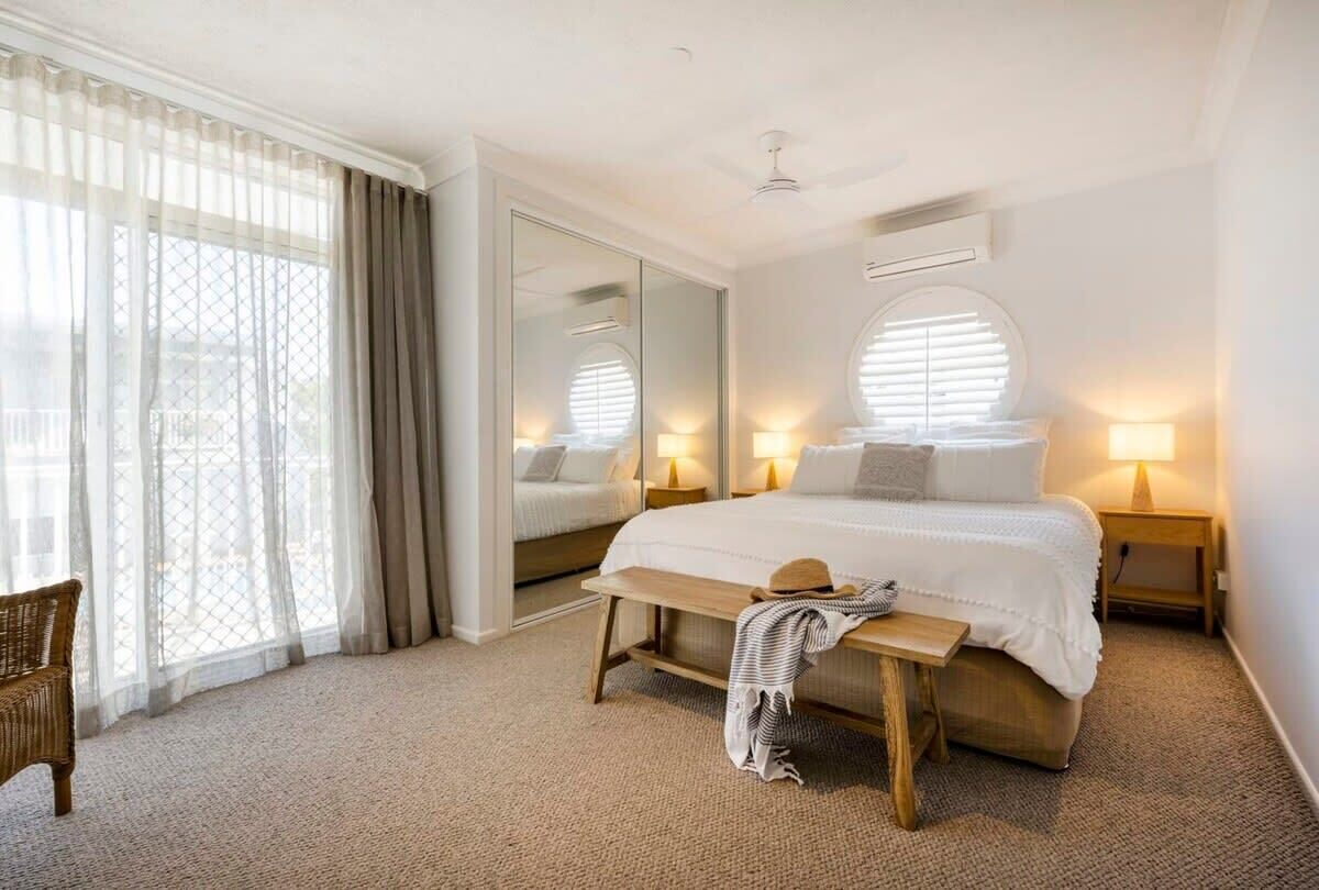 A bedroom with a large bed, bedside lamps, a bench, a mirror, and a view through a window with sheer curtains.