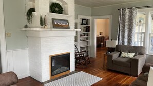 Un salon avec une cheminée en briques blanches, un plancher en bois, un canapé gris et un tapis à motifs.
