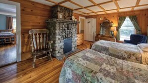 Une chambre confortable, aménagée dans le style d’un chalet, avec des murs en bois et une cheminée en pierre. On y trouve deux lits avec une literie à motifs floraux et un fauteuil à bascule en bois.