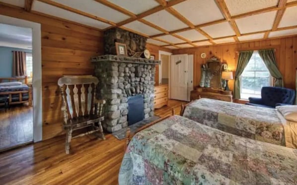 A cozy cabin-style room with wooden walls and a stone fireplace. There are two beds with floral bedding and a wooden rocking chair.