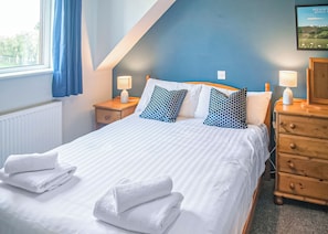 Ein ordentlich bezogenes Bett mit weißen Bettwäsche und blauen Kissen mit Muster, ein Nachttisch aus Holz mit einer Lampe und eine blaue Wand mit einem gerahmten Bild.