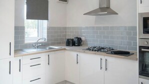 A modern kitchen with white cabinets, a gas stove, a microwave, and a window with a blind. - 2 Bedroom Apartment with Hot Tub (Thirsk)