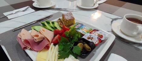 A breakfast setting with a variety of foods including meats, cheese, vegetables, and a dessert on a white plate, accompanied by cups of coffee and tea.