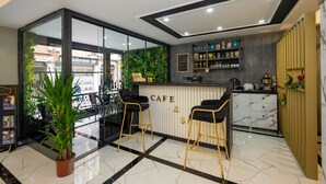A modern cafe with a marble floor, two black bar stools, a counter with a sign reading "CAFE & BAR", and a green wall with plants. - California Hotel (Istanbul)