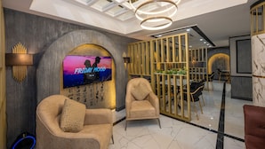 A modern lobby with a large TV screen displaying 'FRIDAY MOOD', two plush armchairs, a dining area with black chairs, and a ceiling with skylights. - California Hotel (Istanbul)