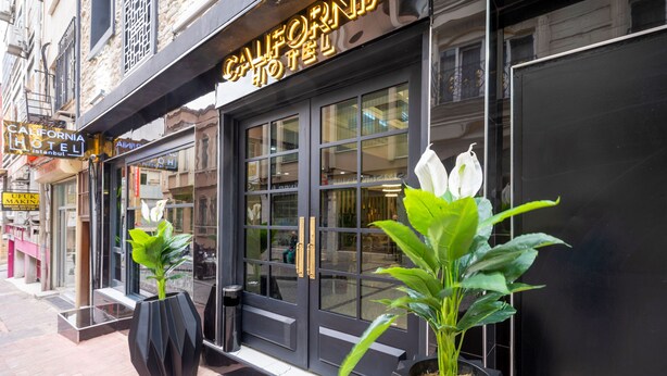 A hotel entrance with a black facade, large glass doors, and two potted plants with white flowers. - California Hotel (Istanbul)