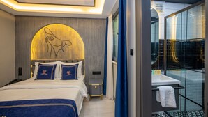 A modern hotel room with a large bed, a small bedside table, a wall-mounted TV, and a bathroom with a glass shower. - California Hotel (Istanbul)