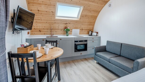 Een compacte keuken met een houten plafond, een eettafel met stoelen, een grijze bank en een klein raam.