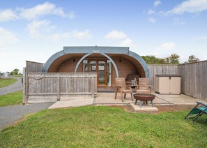 A rustic, semi-circular building with a wooden exterior and a metal roof, featuring a patio with chairs and a table, and a small shed in the background.