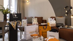 Ein modernes Wohnzimmer mit einem braunen Sofa, einem Couchtisch und einem Bücherregal. Mehrere Luxus-Einkaufstaschen, darunter Chanel und Louis Vuitton, liegen verstreut herum.