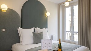 Ein ordentlich bezogenes Bett mit Kissen, ein Nachttisch mit Flasche und Gläsern, eine Geschenktüte und eine Macaron-Schachtel.