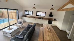 A spacious living area with a black leather sofa, a wooden dining table, and a kitchen with white cabinets and wooden beams. - Wooden villa with pool (Arès)