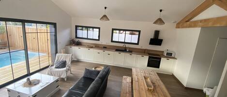 Amplia sala de estar con un sofá de cuero negro, una mesa de comedor de madera y una cocina con armarios blancos y vigas de madera.